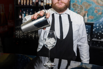 Professional bartender with a beard pours liquid into a glass of ice cubes from a tool for mixing and preparing alcoholic beverages cocktails metal shaker