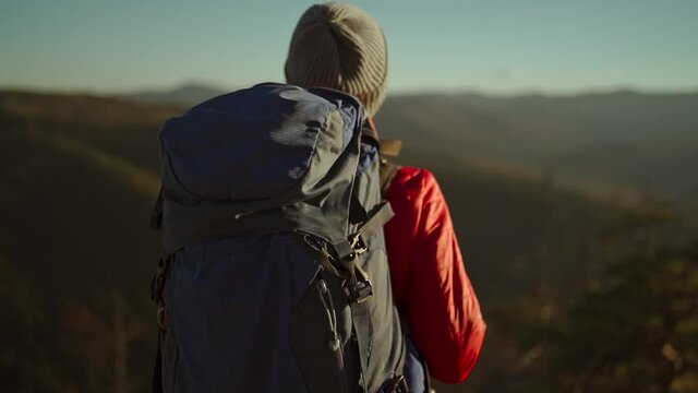 slow motion view from behind adventurous female hiker backpacker in red jacket with backpack stands on mountain top at sunset above the woods.