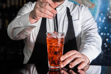 The hand of a professional bartender stirs the red syrup in an alcoholic cocktail with a bar spoon on the bar counter. The process of preparing an alcoholic beverage