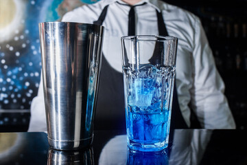 A glass with ice and blue syrup stands on the bar counter against the background of a professional bartender, next to a tool for mixing and preparing alcoholic cocktails with a metal shaker