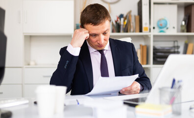 Fototapeta premium Pensive adult businessman sitting at office desk, working with papers..