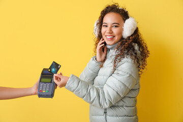 Young woman in winter clothes making payment via banking terminal on color background