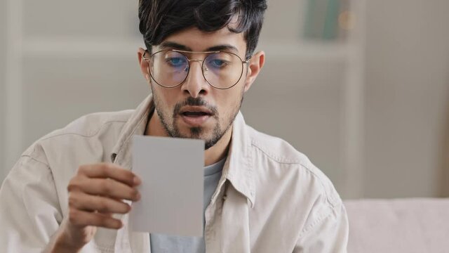 Portrait young arabic man bearded ecstatic guy with glasses sits at home indoors receives envelope letter postcard with good news offer winning victory triumph congratulations feeling happiness joy