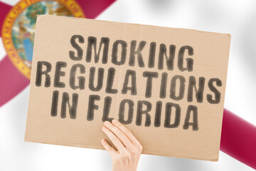 The phrase " Smoking Regulations in Florida " on a banner in men's hand with blurred Florida flag on the background. Judicial. Outside. Place. Pollution. Unlawful. Denial. Denied