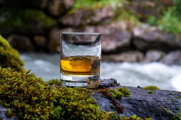 Fotobehang Glass of strong scotch single malt whisky with fast flowing mountain river on background © barmalini