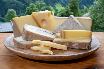 Cheese collection, wooden board with French cheeses comte, beaufort, abondance, emmental, morbier and french mountains village in Haute-Savoie on background