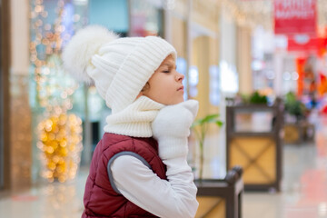 surprised boy in white bobble hat and scarf at mall near showcase. blurred bright lights on background