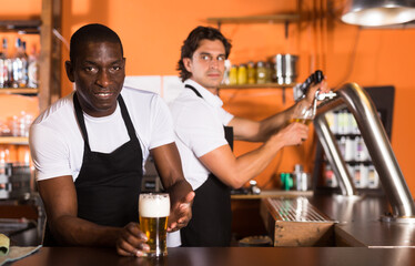 Smiling man bartender offering glass of golden beer, man pouring beer on background in bar