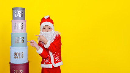 Fototapeta na wymiar A child points with his index fingers to a tower made of round gift boxes with the inscriptions New Year 2022, dressed in Sanat claus clothes, with an artificial beard, copy space.