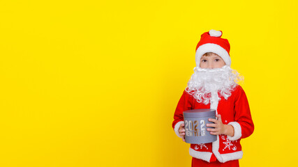 Fototapeta na wymiar Medium portrait of a cute child boy in Santa Claus clothes and with an artificial beard, who is holding a round gift box with the words New Year 2022, yellow, copy space.