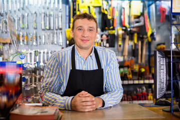 Positive cheerful smiling seller standing in hardware shop - obrazy, fototapety, plakaty