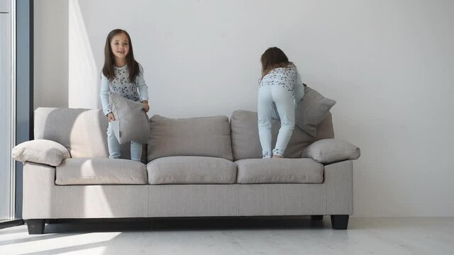 Jumping with pillow on the bed together. Two cute little girls in nightwear at daytime in modern room