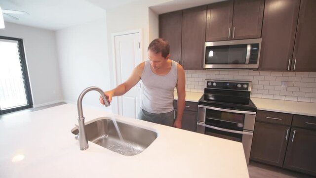 Man checking inspecting stainless steel sink faucet, turning on off tap running water at kitchen room island with quartz countertop at modern apartment by electric microwave oven and brown cabinets