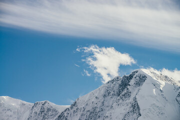 Great view to high snowy mountain peaked top with low cloud under cirrus clouds in sky. Low clouds on big snow covered mountains with sharp pinnacle in sunshine. White-snow pointy peak in sunlight.