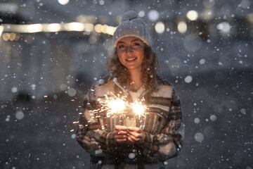 Night portrait of beautiful smiling caucasian woman 25-30 years old in fashion coat and knitted hat celebrating new year party at street with sparklers bengal lights on winter holidays outdoor