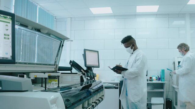 A diverse team works in the lab using tablet computers, wearing masks and gowns. Black male laboratory assistant watching how robot works. Work in the laboratory. High quality 4k footage