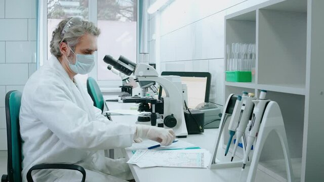 Laboratory assistant works with a microscope and writes notes at white lab. An adult male is working in laboratory with test tubes wearing mask. High quality 4k footage