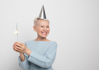 Portrait of a senior woman in studio on a gray background. Woman wearing blue sweater and party hat, holding sparkles.