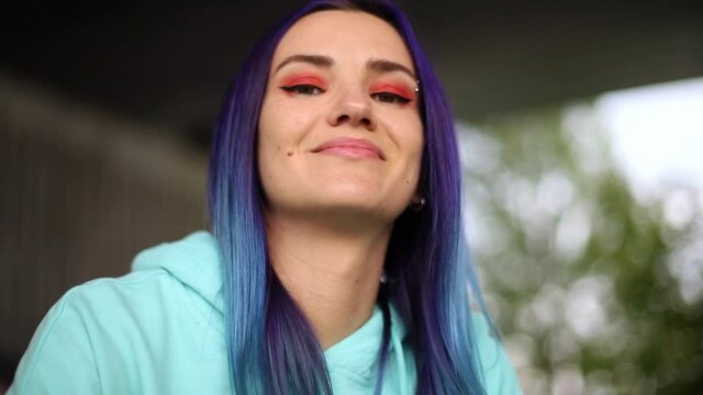 portrait of an ayytacive blue-haired woman with bright make up and brow piercing fixing her hair, smiling and looking to the camera