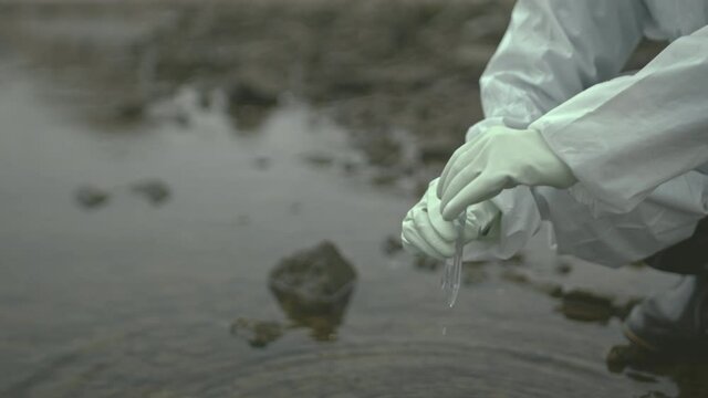 Slowmo close up of unrecognizable ecologist in protective coverall and gloves taking sample of water in polluted river or lake with test tube