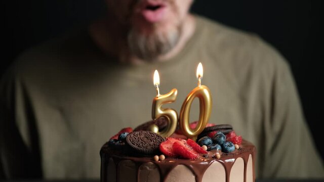 Fifty year old man blows out burning numbers 50 candles on chocolate cake. Birthday cake for 50 years anniversary. Slow Motion