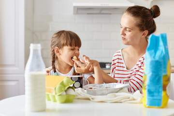 Horizontal shot of happy woman sitting at table with her daughter and eating tasty cakes, posing in kitchen, enjoying homemade pastry, expressing positive emotions.