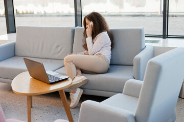 Young millennial woman sitting on a sofa with laptop and talking on mobile phone