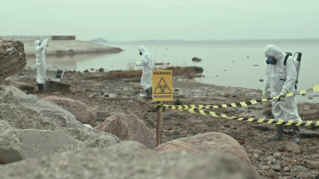 Slowmo shot of team of three scientists in chemical suits exploring dangerous area with yellow biohazard sign near seashore