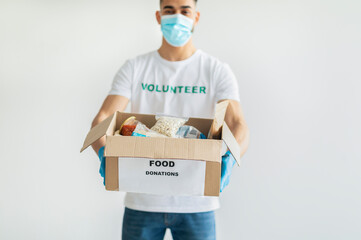 Food donations. Millennial arab man in volunteer t-shirt and medical mask, showing box with canned food and packages