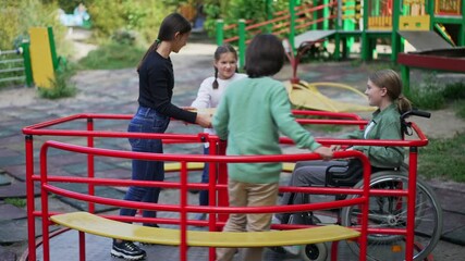 Group of Caucasian children enjoying leisure on playground with friend in wheelchair. Positive boys and girls talking spinning on merry go round smiling. Friendship and disability concept