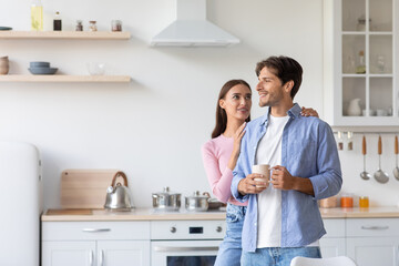 Cheerful millennial husband with cup of hot drink looks at empty space, wife hugs guy in white kitchen