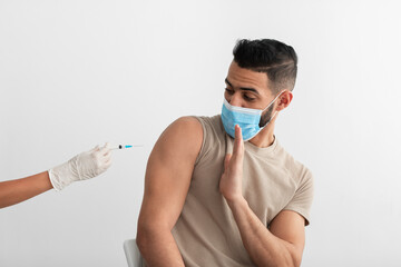 Young Arab guy in disposable mask stopping doctor from making coronavirus vaccine shot, being...