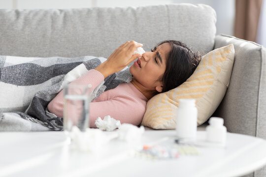 Despaired upset millennial indian woman covered with blanket, suffers from flu blows nose in napkin