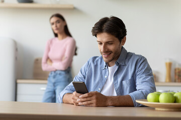 Unhappy upset offended young wife offended at husband with smartphone at kitchen interior