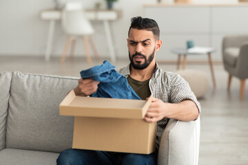 Bad delivery service. Angry Arab man unpacking cardboard box, receiving wrong item, feeling upset with parcel at home - obrazy, fototapety, plakaty