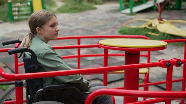 Lonely sad disabled Caucasian girl in wheelchair spinning on merry go round in slow motion. Portrait of devastated child on playground outdoors alone. Disability and childhood concept