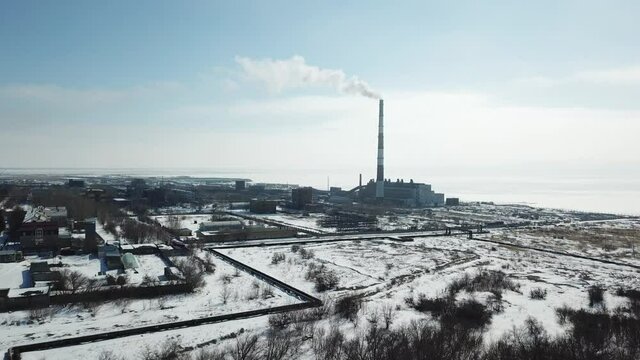Forward moving aerial shot to snowed heat and power plant and its smoking chimney when weather is sunny. Iced big water behind plant on the right of frame, some buildings on the left. Blue sky