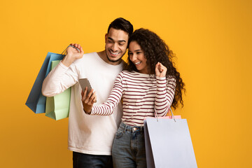 Cashback. Joyful Arab Couple Holding Shopping Bags And Celebrating Success With Smartphone