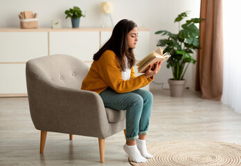 Free time to relax. Young calm arab woman sitting on comfortable armchair and reading paper book at home