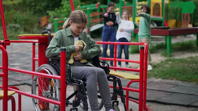 Crying desperate Caucasian girl in wheelchair hugging toy as blurred children pointing laughing at background. Portrait of stressed bullied disabled child on playground with rude kids. Slow motion