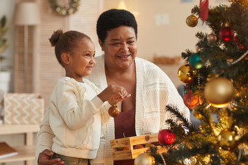 Portrait of cute African-American girl decorating Christmas tree with grandmother while enjoying holiday season at home in cozy interior