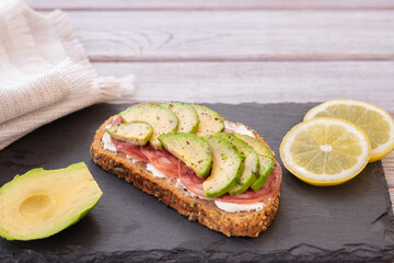 A slice of whole grain bread with seeds with cheese, salami  and sliced avocado for a healthy and tasty snack on a black cutting board. Copy space