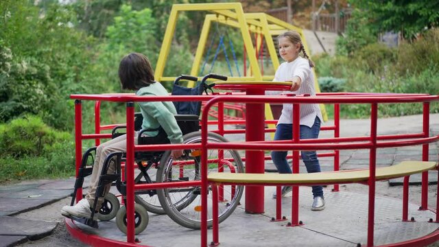 Wide shot positive pretty girl spinning merry go round with disabled cute boy in wheelchair. Charming Caucasian child playing with paralyzed friend on playground in park outdoors. Slow motion