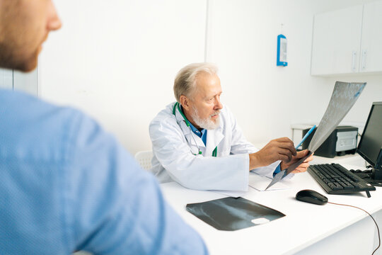 Close-up view from back of young male patient listening mature adult doctor explaining results of MRI scanning, giving professional consultation for treatment illness, explaining causes of disease.