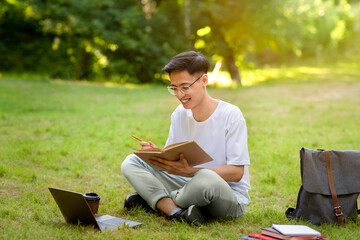 Outdoor Learning. Smiling Asian Male Student Reading Book On Lawn In Park