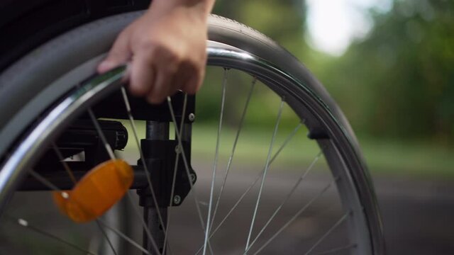 Close-up child hand rolling wheel of wheelchair in slow motion leaving. Unrecognizable Caucasian little disabled boy riding manual assistive device outdoors in summer park
