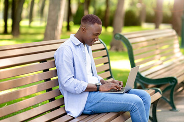 Fototapeta premium Portrait of cool black guy using laptop on bench at park, working remotely, communicating on webcam