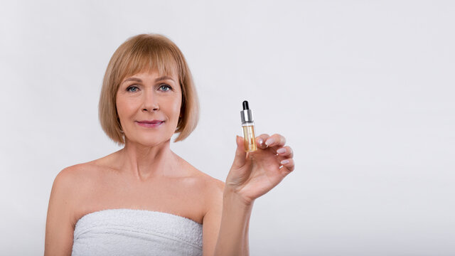 Attractive mature lady with healthy shining skin holding bottle of moisturizing serum on light background, panorama