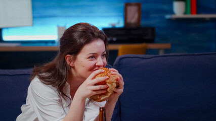 Young person laughing at TV while eating hamburger and drinking beer from bottle on couch. Woman...