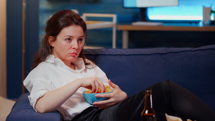 Caucasian woman laying on couch and eating snack while watching television at home. Young adult...